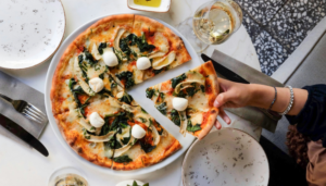 A freshly baked pizza topped with spinach, mozzarella balls, and vegetables on a white plate, with a hand picking up a slice from the table setting.