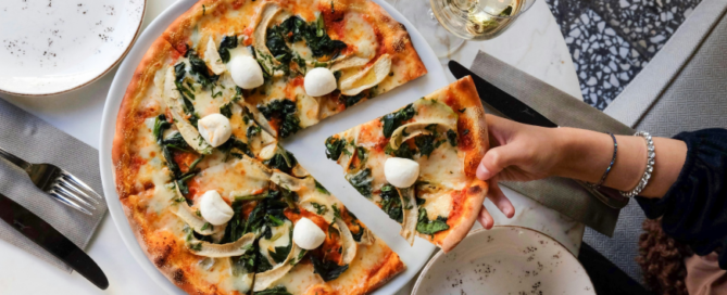 A freshly baked pizza topped with spinach, mozzarella balls, and vegetables on a white plate, with a hand picking up a slice from the table setting.