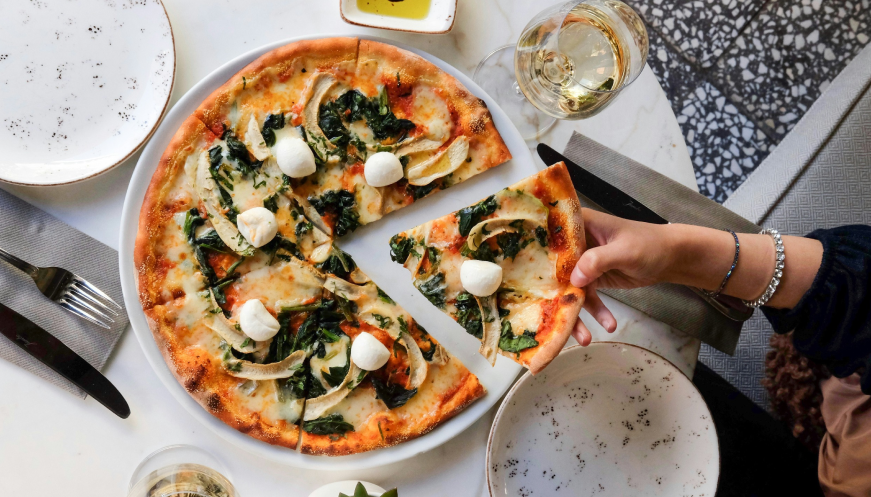A freshly baked pizza topped with spinach, mozzarella balls, and vegetables on a white plate, with a hand picking up a slice from the table setting.
